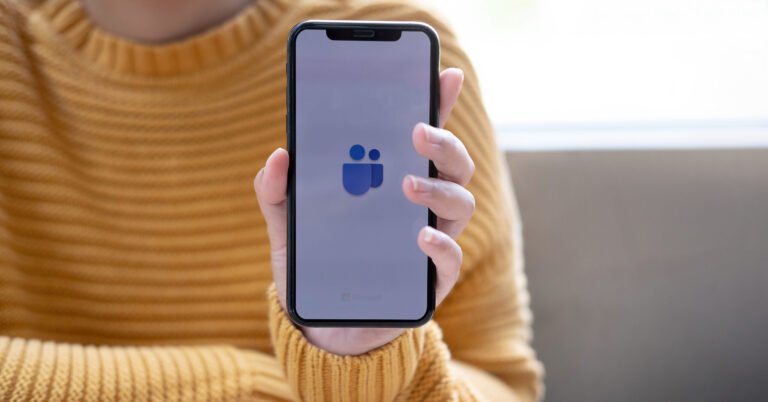 Osoba v žltom svetri drží smartfón s logom Microsoft Teams na obrazovke.