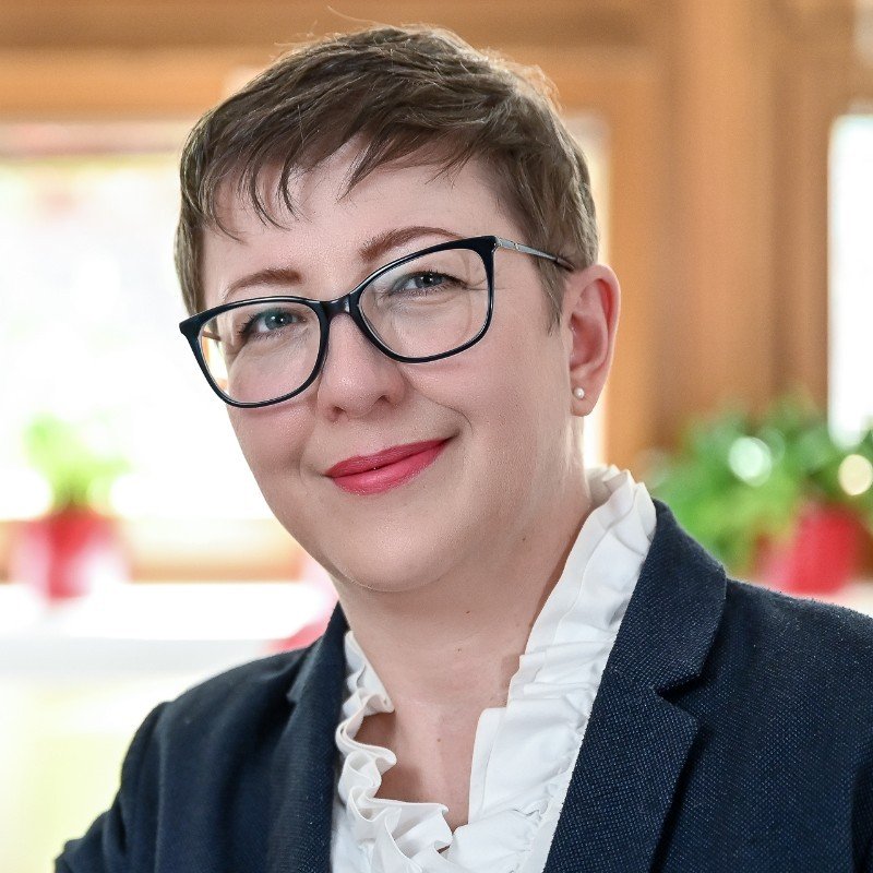 Osoba s krátkymi hnedými vlasmi, v okuliaroch, námorníckom saku a bielej volánovej blúzke sa usmieva do kamery. V pozadí sú mäkko zaostrené izbové rastliny a teplé osvetlenie.