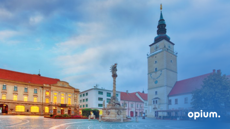 Európske námestie za súmraku s historickými budovami, vysokou hodinovou vežou a centrálnym kamenným pamätníkom. V pravom dolnom rohu je bielym textom cez modrý gradient zobrazené slovo opium.