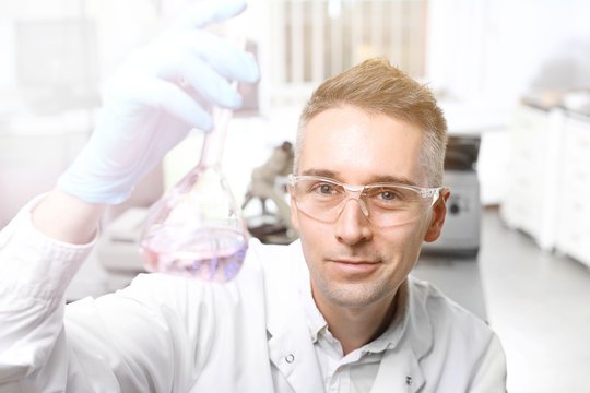 Vedec v ochranných okuliaroch a rukaviciach drží v ruke banku s fialovou tekutinou a v jasne osvetlenom laboratóriu si ju pozorne prezerá.